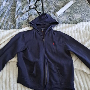Marinblå hoodie från Polo Ralph Lauren - Marinblå hoodie från Polo Ralph Lauren i storlek XL. Tröjan har dragkedja framtill, huva och kängurufickor. Klassisk röd broderad logga på bröstet. Perfekt för chill dagar och enkel att matcha med jeans eller joggers.