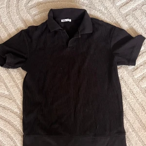 Svart ribbad pikétröja från Zara - Svart pikétröja från Zara i storlek S. Tröjan har korta ärmar, klassisk krage och är ribbad för en snygg struktur. Perfekt för en clean och stilren look. Materialet känns mjukt och bekvämt.
