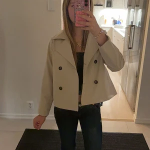 Beige croppad trenchcoat - Snygg beige croppad trenchcoat med dubbelknäppning och bred krage. Jackan har långa ärmar och svarta knappar framtill. Aldrig typ använt den