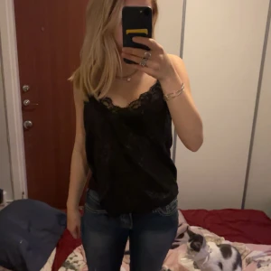 Svart glittrig linnetopp Rut&Circle - Säljer en svart linnetopp från Rut&Circle med tunna axelband och spetsdetaljer vid urringningen. Toppen har ett glittrigt tyg som ger en snygg effekt och är perfekt för dig som vill sticka ut. Sitter löst och har en feminin v-ringning.