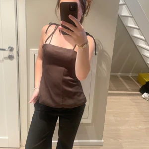 Brun linnetopp med knytband - Säljer en brun linnetopp med smala axelband som knyts på axlarna. Toppen har en rak, något figurnära passform och är tillverkad i ett slätt material som ger en stilren look. Perfekt för dig som gillar minimalistisk stil och vill ha något enkelt men snyggt.