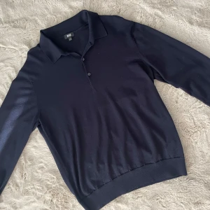 Mörkblå stickad pikétröja från Uniqlo - Snygg mörkblå stickad pikétröja från Uniqlo med klassisk krage och knäppning framtill. Tröjan har lång ärm och ribbade muddar vid ärmslut och nederkant. Perfekt för dig som gillar stilrena och bekväma plagg.