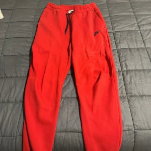 Röda mjukisbyxor från Nike, storlek M - Röda mjukisbyxor från Nike med svart logga på vänster lår. Byxorna har resår i midjan med svart snörning och är tillverkade i mjukt material. Perfekta för chill eller träning, med klassisk sportig look och bekväm passform.
