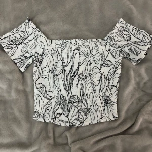 Offshoulder blommig topp från H&M - Snygg offshoulder topp från H&M i vit och svart med blommigt mönster. Gjord i linnemix för en luftig känsla. Toppen har korta ärmar och smockad struktur som ger en cool och trendig look.
