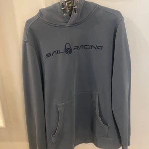 Blå hoodie från Sail Racing  - Blå hoodie från Sail Racing med broderad logga på bröstet och emblem på huvan. Tröjan har snörning i halsen, stor ficka på framsidan och är tillverkad i mjuk bomull. Perfekt för chill dagar och snygg till jeans eller joggers. Storlek M