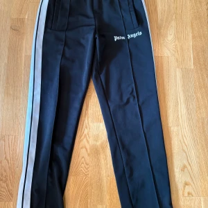Palm Angels svarta trackpants M - Svarta trackpants från Palm Angels med vita sidoränder och logga tryckt på låret. Byxorna har elastisk midja och är tillverkade i polyester för en sportig vibe. Perfekta för en streetwear-look.