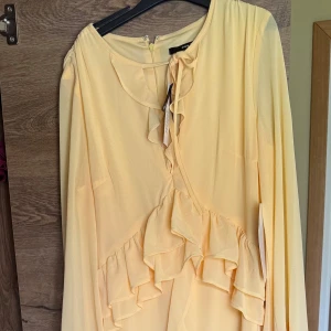 Ljusgul omlottklänning med volanger - Supersöt ljusgul omlottklänning från Asos med långa ärmar och volangdetaljer framtill. Klänningen har en feminin v-ringning och knyt i halsen. Perfekt för dig som gillar romantisk och trendig stil.