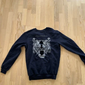 Mörkblå sweatshirt Kings Will Dream XS - Mörkblå sweatshirt från Kings Will Dream i storlek XS. Tröjan har ett stort tryck av ett tigerhuvud i grått och vitt framtill samt ribbade muddar vid ärmslut och nederkant. Perfekt för dig som gillar streetstyle och snygga prints.