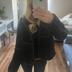 Svart teddyjacka Nelly - Säljer en svart teddyjacka med loose passform och markerade sömmar. Jackan har en bred krage, knappar framtill och är långärmad. Materialet är mjukt och fluffigt, perfekt för dig som gillar chill och comfy vibes.