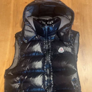 Monclerväst - Svart, glansig dunväst från Moncler med avtagbar huva och ikonisk logga på bröstet. Västen har dragkedja med Moncler-berlock och quiltad design. Insidan har ett coolt serietidningsmönster och märkning för äkthet. Perfekt för att höja din streetstyle. Köpt för 3500kr får även med massa tags och en påse
