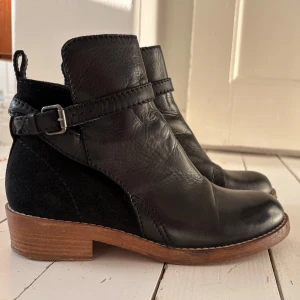 Svarta boots från Acne Studios - Snygga svarta boots från Acne Studios i storlek 38. Skorna har en kombination av skinn och mocka, med dekorativ rem och spänne runt ankeln. Brun sula med låg klack ger en stilren kontrast. Perfekta för dig som gillar minimalistisk och trendig design.