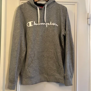 Grå hoodie från Champion med logga - Grå hoodie från Champion med vit logga framtill och vita snören i huvan. Klassisk känguruficka och mjukt material som passar perfekt till chill eller träning. Långärmad och normal passform, enkel att matcha med jeans eller joggers.