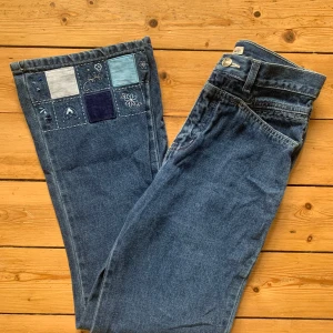 Blå jeans med patchwork och broderi W26 L28 - Unika blå jeans från Authentic med patchwork och broderade detaljer i olika blå och vita nyanser längst ner på benen. Jeansen har raka ben och klassisk femficksdesign. Materialet är robust denim i bomull, perfekt för dig som vill sticka ut med en kreativ look.