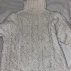 Vit kabelstickad polotröja - Mysig vit kabelstickad polotröja med hög krage och grova flätmönster. Perfekt för kalla dagar och ger en klassisk vibe. Tröjan har lång ärm och är riktigt chunky i modellen.