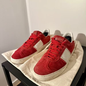 Valentino Garavani röda sneakers mocka - Riktigt snygga Valentino Garavani sneakers i röd mocka med vita skinnpaneler och röda skosnören. Klassisk låg modell med rund tå och platt sula. Snygga nitar på hälen ger extra edge. 1 nit saknas på högra skon men det går att få med extra nitar (OBS! Extra nitarna är ej äkta utan kopior) bara att höra av sig vid funderingar👍