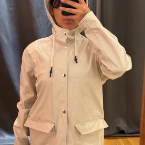 Beige regnjacka från 157 med huva - Beige regnjacka från 157 med huva och dragsko, stora fickor med lock och tryckknappar framtill. Jackan har en rak passform och är tillverkad i ett slätt, vattenavvisande material. Perfekt för regniga dagar och enkel att styla till jeans.