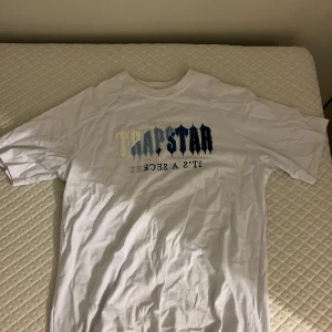 Vit Trapstar t-shirt med broderi - Vit t-shirt från Trapstar, det är för storlek L, men står M, med broderad logga i olika blå och beige toner på bröstet. Klassisk passform med rund hals och korta ärmar. Materialet är mjuk bomull och trycket har en tydlig streetkänsla. Perfekt för dig som gillar snygga statement-plagg.