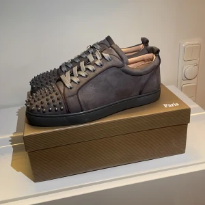 Christian Louboutin skor - Snygga mörkgrå sneakers i mocka med röda sulor och coola silverfärgade nitar på tån. Skorna har snörning och en klassisk låg siluett. Perfekta för dig som vill sticka ut med exklusiv streetstyle.