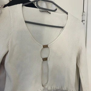 Vit ribbad croppad topp med kedjedetalj - Säljer en vit ribbad croppad topp från Chiquelle med lång ärm och snygga guldfärgade kedjedetaljer framtill. Toppen har en djup v-ringning och öppet parti framtill som hålls ihop av två kedjor. Perfekt för dig som vill sticka ut med en trendig look. Den är väl använd därav lite defekter i materialet, den guldiga färgen på detaljerna framme är även inte helt kvar.