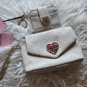 Vit axelväska & plånbok Juicy Couture - Superfin vit axelväska och matchande plånbok från Juicy Couture. Båda har präglat JC-mönster, guldfärgade detaljer och hjärtformade spännen med rosa stenar. Väskan har kedjerem och innerfoder med logga. Perfekt för dig som gillar söta detaljer och lyxig vibe.