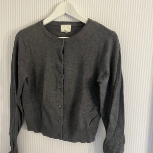 Mörkgrå kofta från H&M, storlek S - En stilren mörkgrå kofta från H&M i storlek S. Koftan har rund halsringning, långa ärmar och knäppning med matchande knappar framtill. Tillverkad i mjuk bomullsblandning som är perfekt för lager-på-lager under höst och vinter.