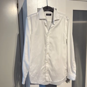 Vit skjorta från Dressmann i bomull - Klassisk vit skjorta från Dressmann i premium bomull med diskret fiskbensmönster. Skjortan har lång ärm, knappar framtill och bred krage. Perfekt för dig som vill ha en clean och stilren look.