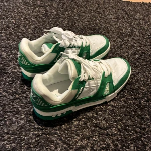 Louis Vuitton skor 43 - Säljer ett par Louis Vuitton sneakers i grönt och vitt med klassiska LV-detaljer. Skorna har snörning, platt sula och rund tå. Materialmix av skinn och syntet med präglade monogram på ovandelen. Perfekta för dig som vill sticka ut med lyxig streetstyle.