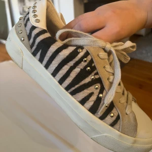 Zebramönstrade sneakers med nitar - Snygga sneakers med zebramönster i svart och beige, dekorerade med silvriga nitar runt hälen och på sidorna. Skorna har snörning och detaljer i mocka och skinn, samt vit sula och tå. Perfekta för dig som vill sticka ut med din stil.