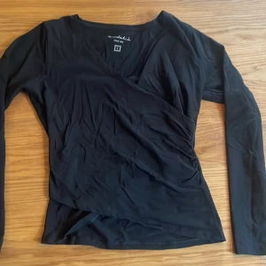 Svart omlottopp från Bondelid - Svart långärmad omlottopp från Bondelid i storlek S. Toppens omlottdesign ger en snygg v-ringning och en smickrande passform. Perfekt att styla med jeans eller kjol för en trendig look.