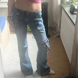 Blå cargojeans med fickor - Säljer ett par blå cargopants i jeansmaterial med flera stora fickor och coola detaljer som spännen och dragkedjor. Byxorna har låg midja och en lite loose passform med raka ben. Perfekta för dig som vill ha något unikt i garderoben. 