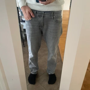 Grå jeansbyxor storlek 32/32 - Säljer ett par grå jeans från Jack & Jones med klassisk femficksdesign och raka ben. Jeansen har en snygg, ljusgrå tvätt och är tillverkade i mjukt jeanstyg. Passformen är normal och midjan är midwaist. Perfekta för en clean och stilren look.       Specifikation.                                                               Jack & Jones chris/relaxed 32/32 