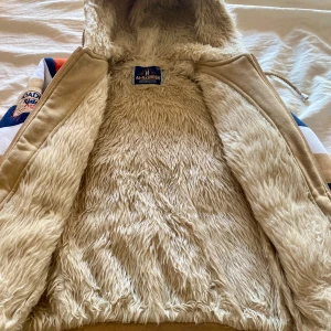 Randig  hoodie med huva  storlek 8-9 år Nytt sikick - Randig hoodie med huva och dragkedja  Jackan har breda ränder i beige, marinblått och vitt, broderade detaljer och patchar framtill. Insidan är helfodrad med mjuk beige fuskpäls. Ribbade muddar med ränder i rött och marinblått.