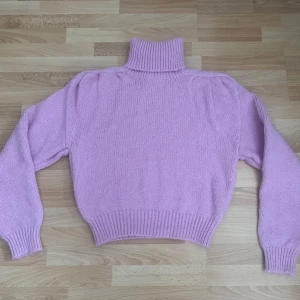 Ljuslila stickad polotröja - Supermysig ljuslila stickad polotröja med puffiga axlar och bred ribbad mudd vid ärmslut och nederkant. Tröjan har en hög polokrage och är tillverkad i en mjuk blandning av akryl, polyester och polyamid. Perfekt för kyliga dagar när du vill vara både varm och snygg. Använd en gång.