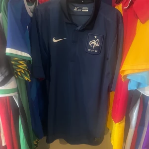 Frankrike Nike fotbollströja marinblå L Äkta - Marinblå officiell Frankrike fotbollströja från Nike med FFF-emblem och vit Nike-logga på bröstet. Tillverkad i Dri-FIT-material som andas, med klassisk krage och korta ärmar. Mycket bra skick, Äkta