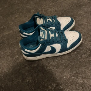 Nike Dunk Low blå/vita sneakers - Nike Dunk Low sneakers i blått/turkos och vitt med klassisk swoosh på sidorna. Ovandelen är i skinn med struktur och perforerad tåbox. Skorna har blå snörning, vit mellansula och blå detaljer på plösen och innersulan. Perfekt för dig som gillar streetwear och snygga detaljer. 