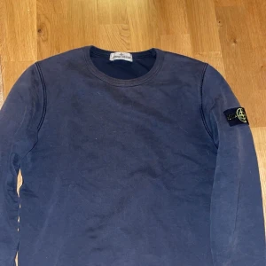 Mörkblå tröja från Stone Island Junior - Snygg mörkblå långärmad tröja från Stone Island Junior med klassisk rund halsringning och ikonisk logotyp-patch på vänster ärm. Tillverkad i mjuk bomull för en skön och avslappnad stil. Perfekt för dig som gillar streetwear och vill sticka ut.