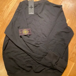 Svart Stone Island sweatshirt - Svart sweatshirt från Stone Island med klassisk rund hals och ribbade muddar. Tröjan har den ikoniska Stone Island-loggan på vänster ärm, fastsatt med knappar. Tillverkad i mjuk bomull för en skön och avslappnad passform.