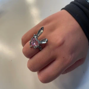 Silverring med rosa hjärta och kaninöron - Unik silverfärgad ring med ett stort rosa hjärtformat stenar i mitten och två kaninöron på toppen. Ringen har små glittriga detaljer runt hjärtat som ger extra bling. Perfekt för dig som gillar söta och annorlunda smycken.