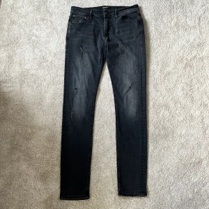 Jack & Jones jeans  - Mörkgrå/svarta slim jeans från Jack & Jones med slitna detaljer och klassisk femficksdesign. Jeansen har en mörk tvätt med lätt urtvättade partier och smal passform hela vägen ner. Perfekta för dig som gillar en modern och stilren look. Storleken är 32/34