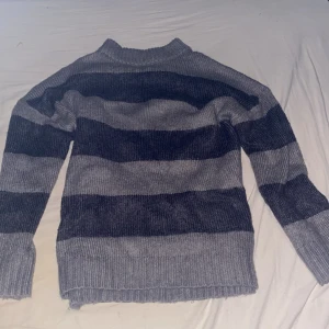 Randig stickad tröja i grå toner - Mysig stickad tröja med breda mörkgrå och ljusgrå ränder. Tröjan har ribbade muddar vid ärmslut och nederkant samt en högre rund halsringning. Perfekt för kyliga dagar och enkel att matcha med jeans eller kjol.