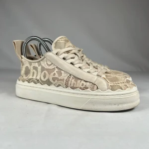 Beige sneakers från Chloé i spets - Unika sneakers från Chloé i beige med snygg spetsdetalj och vågig kant längs sulan. Skorna har snörning och tydlig logga på sidan. Kombination av skinn och spets ger en lyxig och trendig vibe. Perfekta för dig som vill sticka ut med en exklusiv touch.