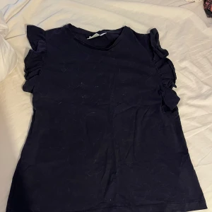 Svart topp med volangärmar - En svart topp med rund halsringning och korta ärmar med volangdetaljer. T-shirten har en enkel, rak passform och är tillverkad i mjukt bomullstyg. Perfekt för dig som gillar en stilren look med en twist på ärmarna.