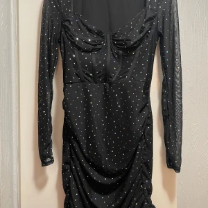 Svart glittrig kortklänning med mesh - Supersnygg svart kortklänning med glittriga prickar och mesh-långärmar. Klänningen har en markerad byst med snygga sömmar och lätt rynkad effekt nedtill. Perfekt för dig som vill sticka ut med lite extra bling och cool siluett.