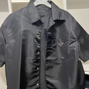 Svart Prada skjorta - Svart skjorta från Prada med korta ärmar och klassisk krage. Skjortan har en bröstficka med Pradas ikoniska metallogo och stängs med tryckknappar. Materialet är glansigt och känns lyxigt, perfekt för dig som vill sticka ut med en clean och modern look. Pris går att diskuteras vid snabb affär tveka inte med frågor                       MVH