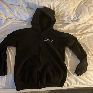 Svart hoodie med dragkedja från Bvaldi - Svart hoodie från Bvaldi i storlek XS med dragkedja och huva. Framsidan har en liten Bvaldi-logga och baksidan är dekorerad med stora silvriga motiv och text. Perfekt för dig som gillar streetstyle och vill sticka ut.