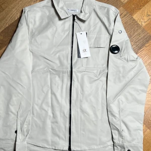 Ljusgrå overshirt från C.P. Company - Stilren ljusgrå overshirt från C.P. Company med klassisk krage, bröstficka och ikonisk linsdetalj på ärmen. Jackan har hel dragkedja framtill och är tillverkad i ett slätt, lätt material. Perfekt för lager-på-lager och snygg till streetwear. Jackan har en medium bredd med närmare s i längd. Skriv gärna om du har frågor
