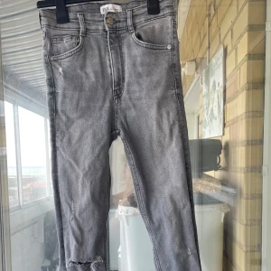 Grå skinny jeans från Zara - Säljer ett par grå skinny jeans från Zara med slitningar och råa kanter vid knäet. Jeansen har klassisk femficksdesign, knapp och dragkedja framtill.