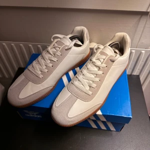 Vita sneakers från Sandö strl 42 - Snygga vita sneakers från Sandö i storlek 42. Skorna har en stilren design med ovandel i vitt skinn och mockadetaljer, samt en klassisk gummisula i brunt. Perfekta för dig som gillar minimalistisk och clean stil. Skorna har snörning och rund tå. Endast provade