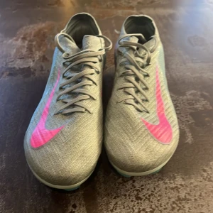Nike fotbollsskor grå med rosa detaljer - Säljer ett par Nike mecurial elite fotbollsskor i grått med stora rosa swoosh-loggor och coola turkosa dobbar undertill. Skorna har snörning och är tillverkade i syntetmaterial för bästa grepp och känsla på planen. Perfekta för dig som vill sticka ut med stil på matchen! Storlek 42 