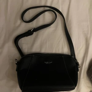 Svart axelväska från Jennifer Jones - Snygg svart axelväska från Jennifer Jones med justerbar rem och silverfärgade detaljer. Väskan är tillverkad i skinnimitation och har dragkedja upptill. Perfekt storlek för det viktigaste och stilren design med vita sömmar.